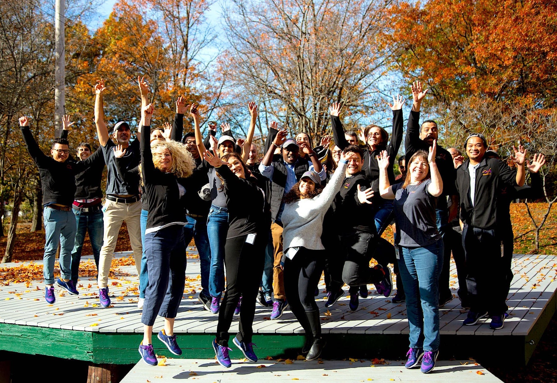 group of people outside surrounded by trees & fall leaves, jumping with hands in the air